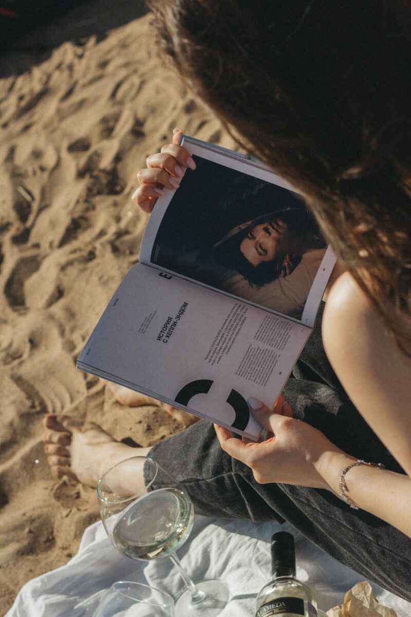 Woman enjoying a sunny day at the beach, reading a magazine with wine at her side.